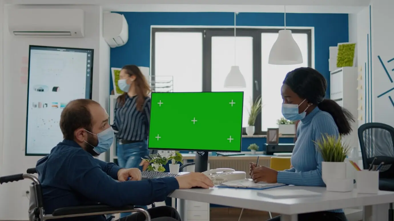 Ein behinderter, gelähmter, invalider Unternehmer mit Gesichtsmaske erklärt einem schwarzen Mitarbeiter die finanzielle Entwicklung des Unternehmens und zeigt dabei auf einen Greenscreen, ein Mock-up und einen Chroma-Key-isolierten Desktop in einem New-Normal-Büro.